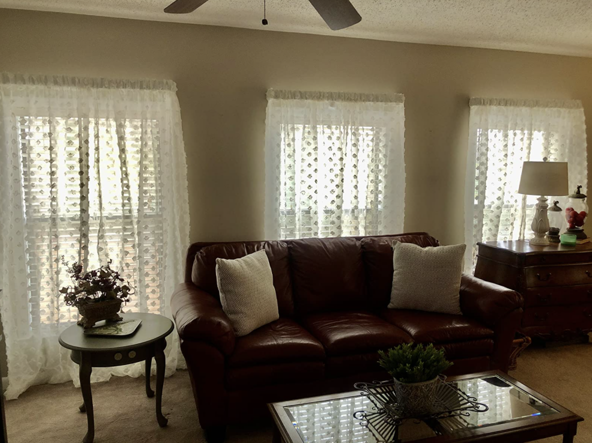 Reviewer&#x27;s full length living room windows covered with white embroidered sheer curtains that let the light in