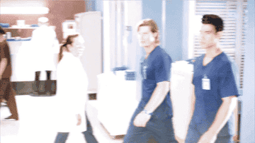GIF of two actors from Grey&#x27;s Anatomy in scrubs and walking through the hospital