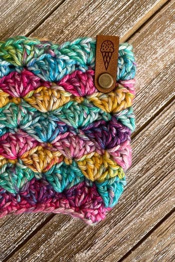 a rainbow crochet pint cozy with a leather tab featuring an engraving of an ice cream cone 
