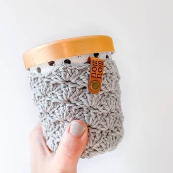 a hand holding up a pint of ice cream with a light gray crochet cozy on it 
