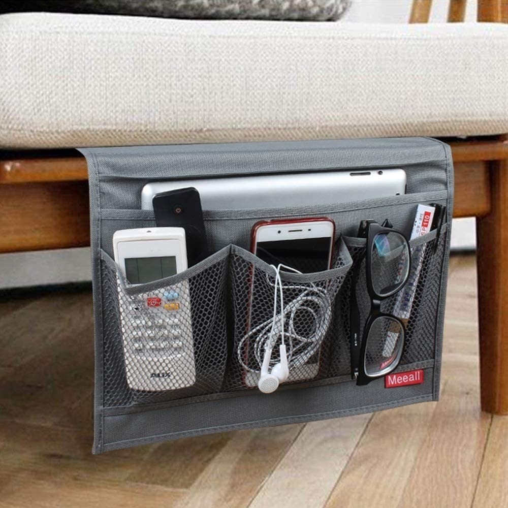 A gray fabric caddy tucked under a mattress with pockets holding a phone, remotes, glasses, and a laptop