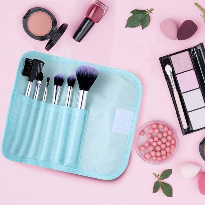 A folding bag in turquoise colour with 7 different makeup brushes placed inside, kept alongside makeup products