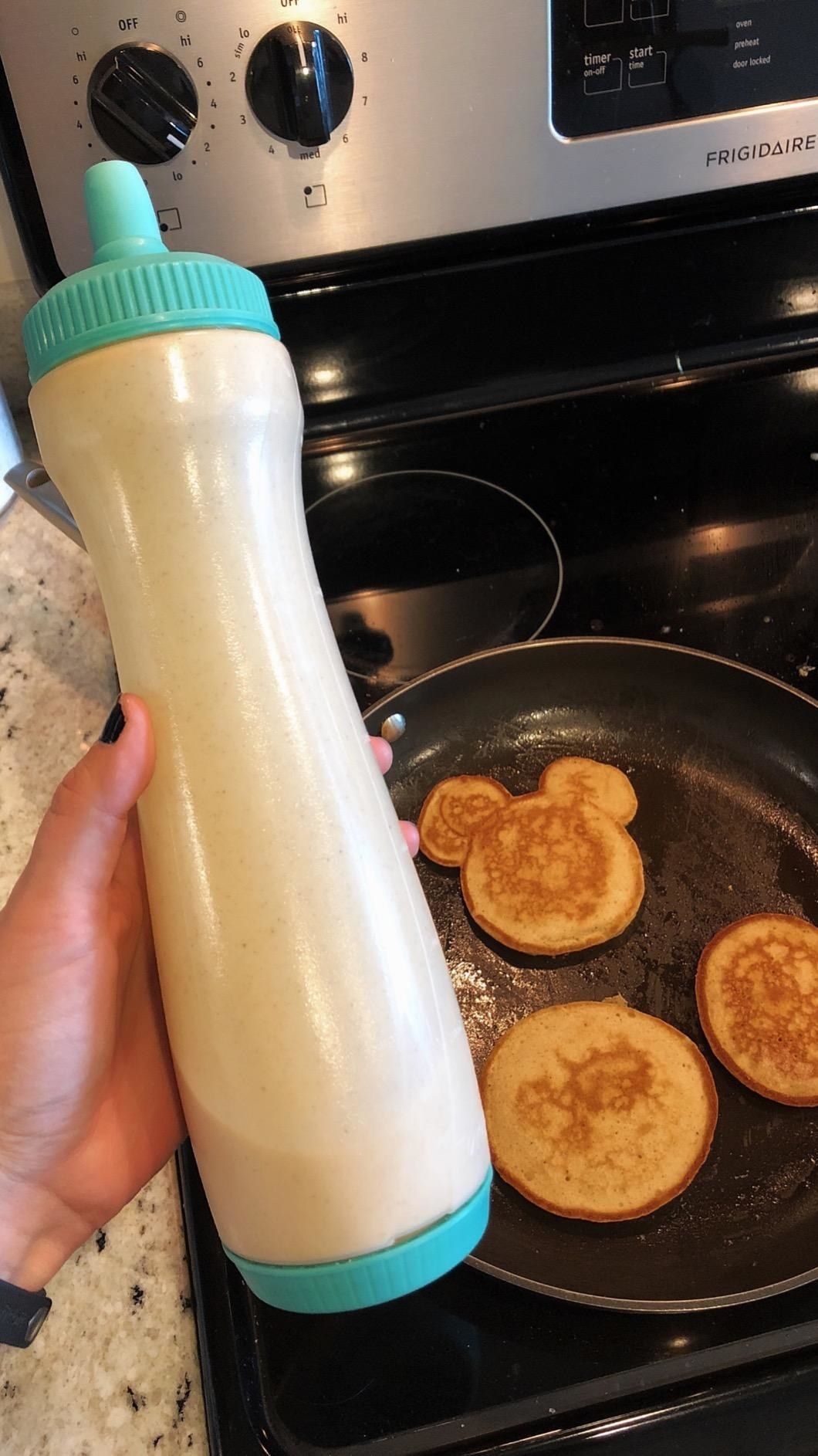image of reviewer holding a full bottle dispenser above mickey mouse shaped pancakes in a pan