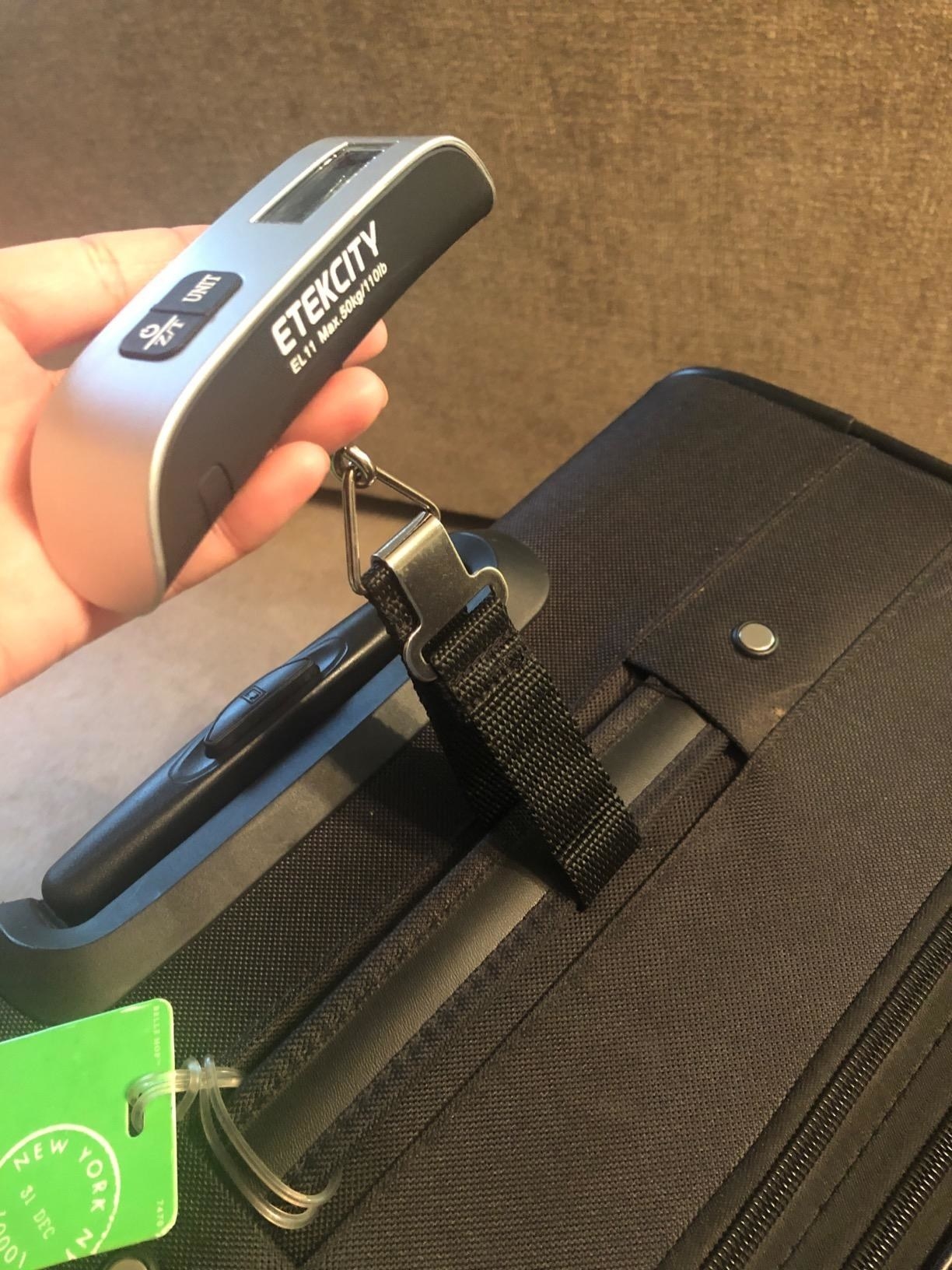 reviewer holding the silver and black luggage scale, attached the the top of a suitcase
