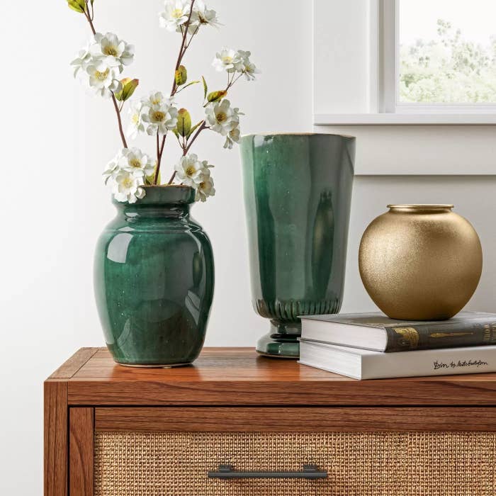 The round brass vase is atop two books and next to green glazed pottery all on top of a brown dresser