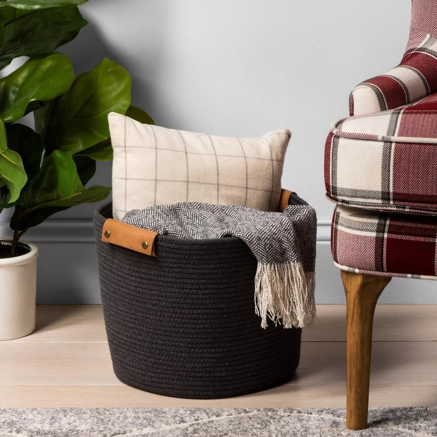 The black coiled basket is holding a pillow and a blanket and is placed next to a chair, rug and large plant