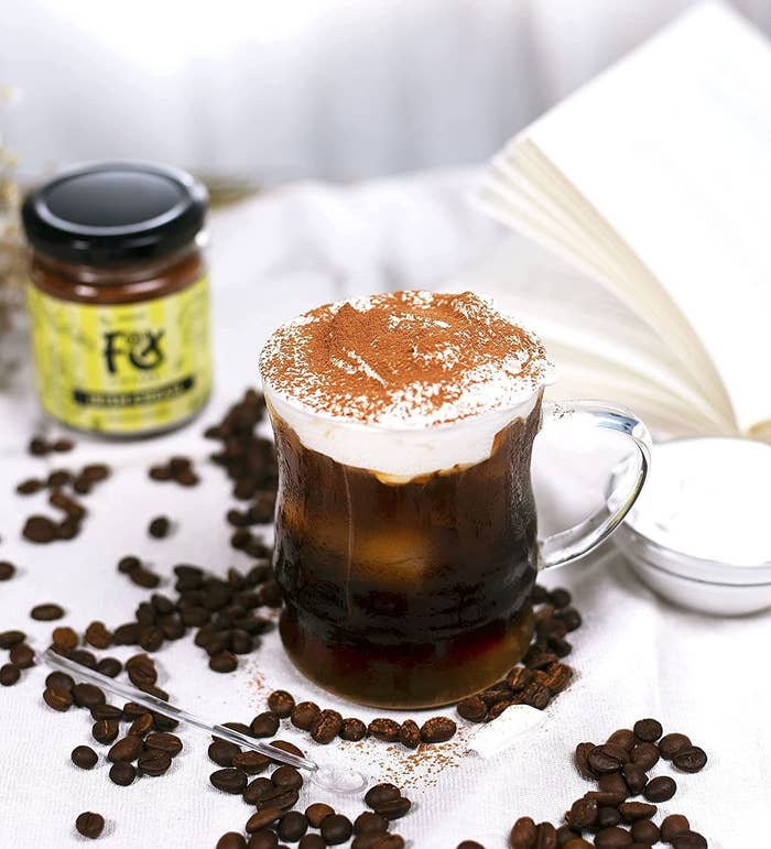 A mug of coffee with whipped cream on top. Coffee beans are scattered around it and the jar of coffee is kept in the background.