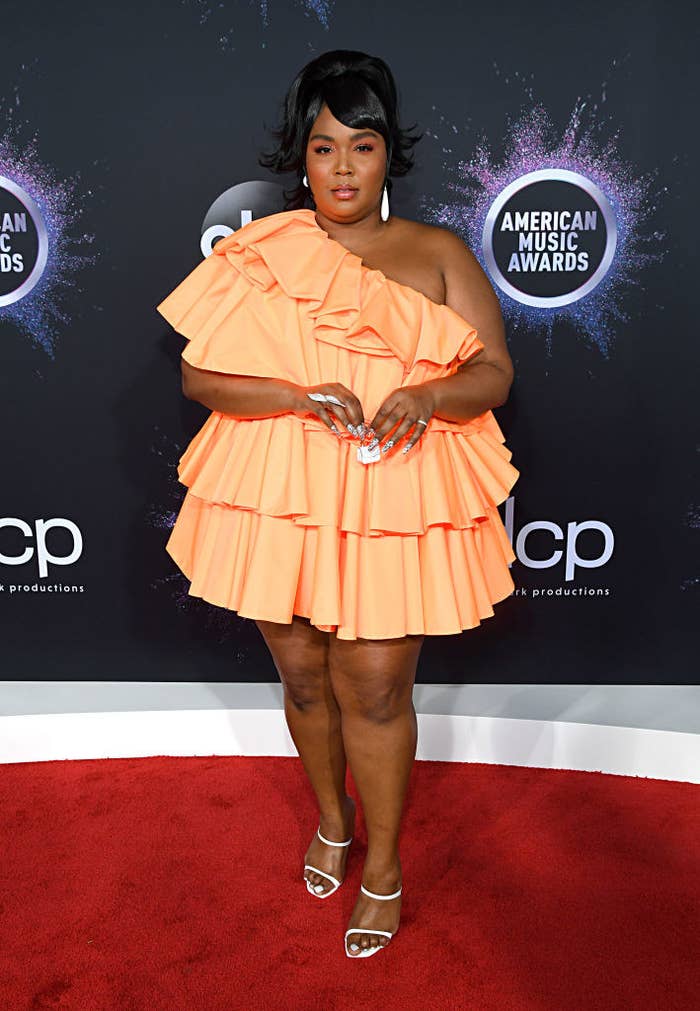 Lizzo posing in a one-shoulder short dress and holding a tiny purse on a red carpet