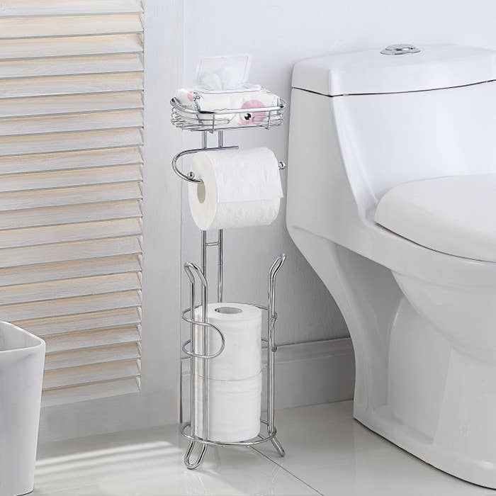 A metallic freestanding toilet paper holder with a space at the top for a cell phone, a space in the middle for toilet paper, and a space below to hold up to three toilet paper rolls