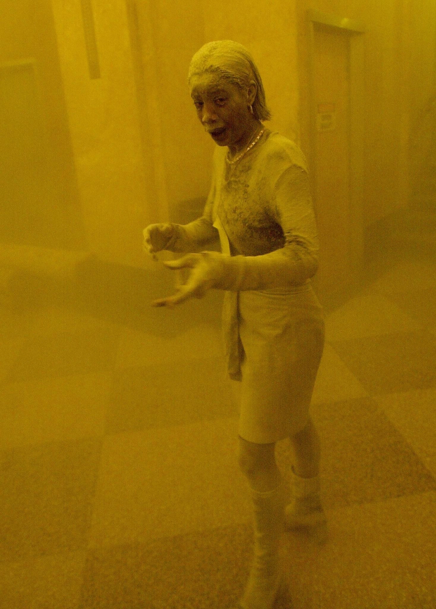 A woman completely covered in ash and in apparent distress in a building lobby that is yellow and hazy from the ash and smoke