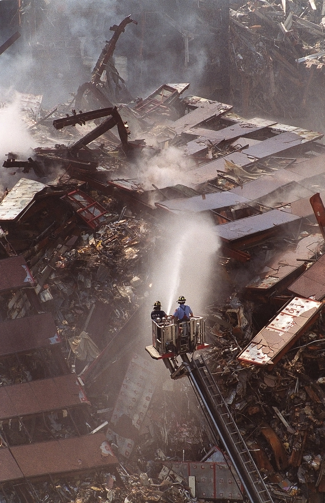 Two firefighters on a platform crane above a mountain of wreckage