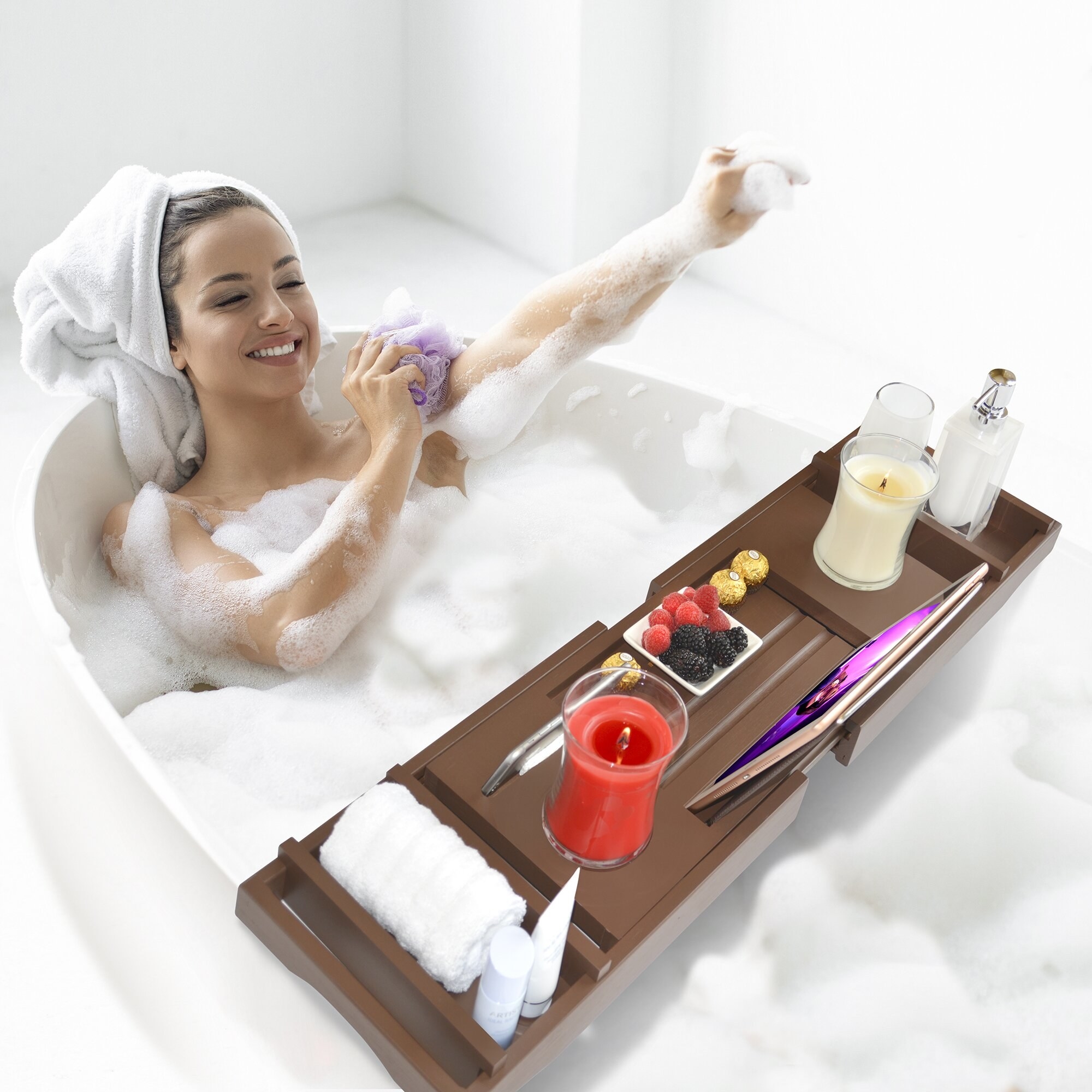 A woman in the bathtub with a caddy board over the tub, with a candle, food, and an electronic tablet