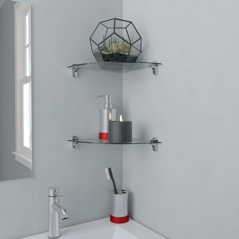 Two glass corner shelves in a gray bathroom. One shelf has soap and a candle on it; the other has a knickknack