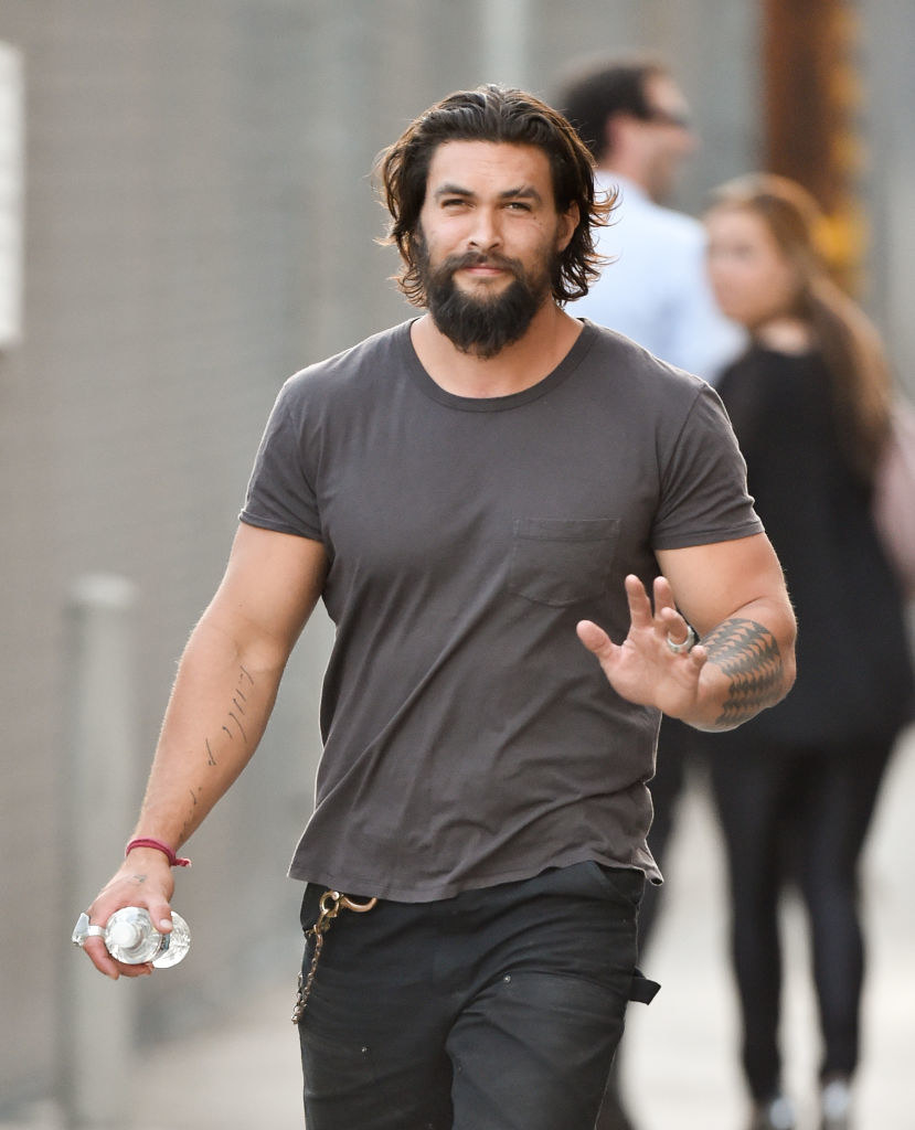 Jason Momoa is seen in a grey top and black pants holding a bottle of water