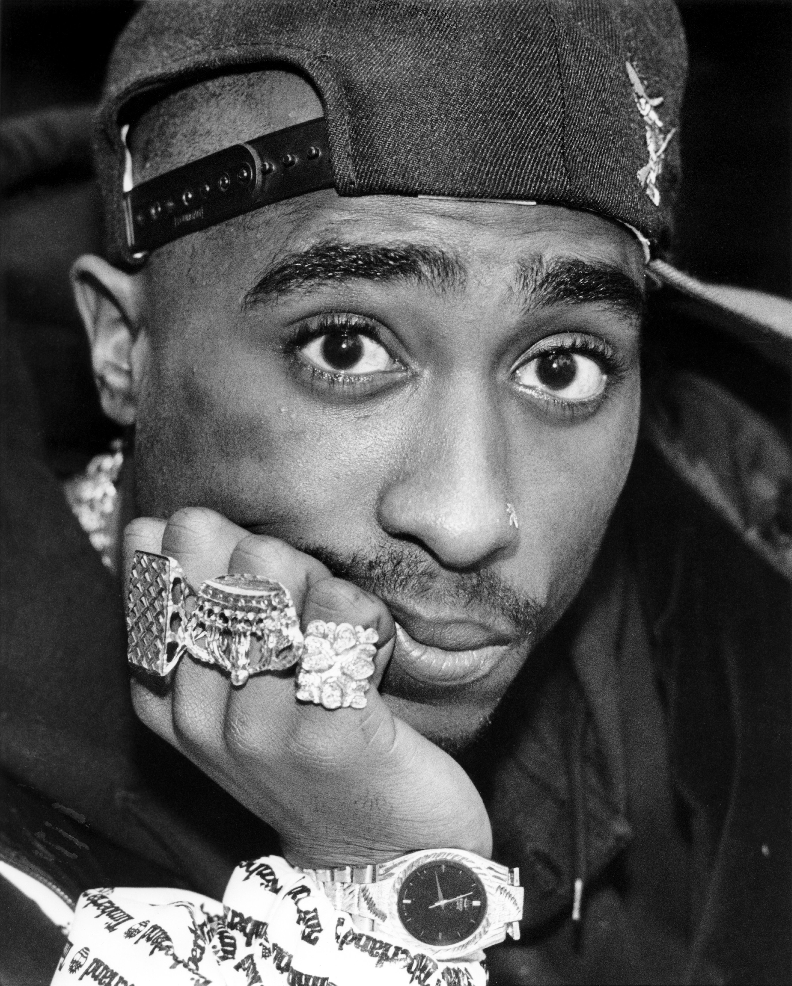 A close-up of Tupac&#x27;s face, wearing a backward cap, rings, and a wristwatch