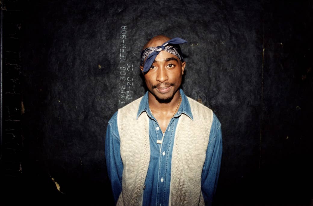 Tupac smiles and wears a bandana, knit vest, and denim shirt