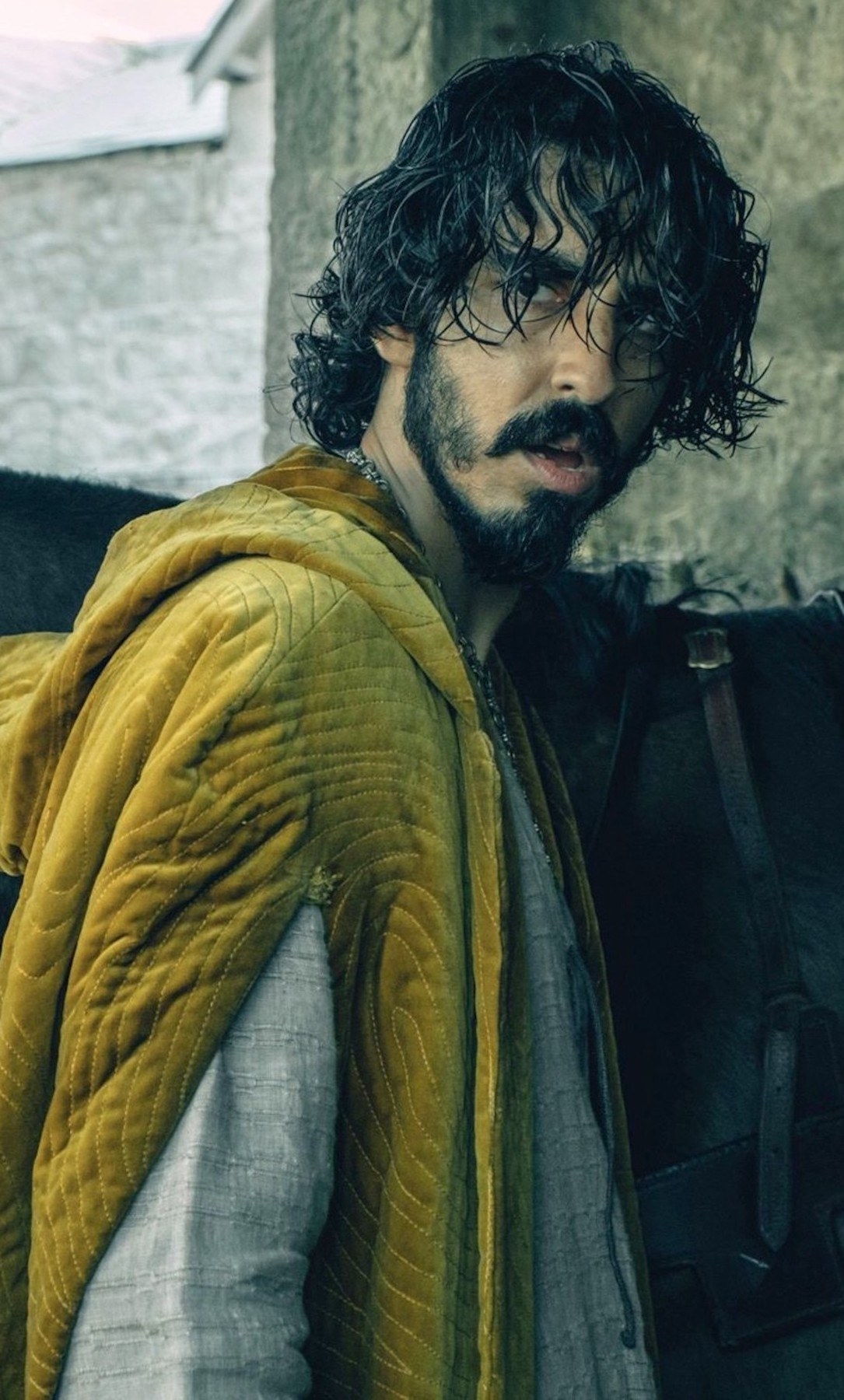 Dev Patel with long hair, a beard, standing next to a horse looking scruffy