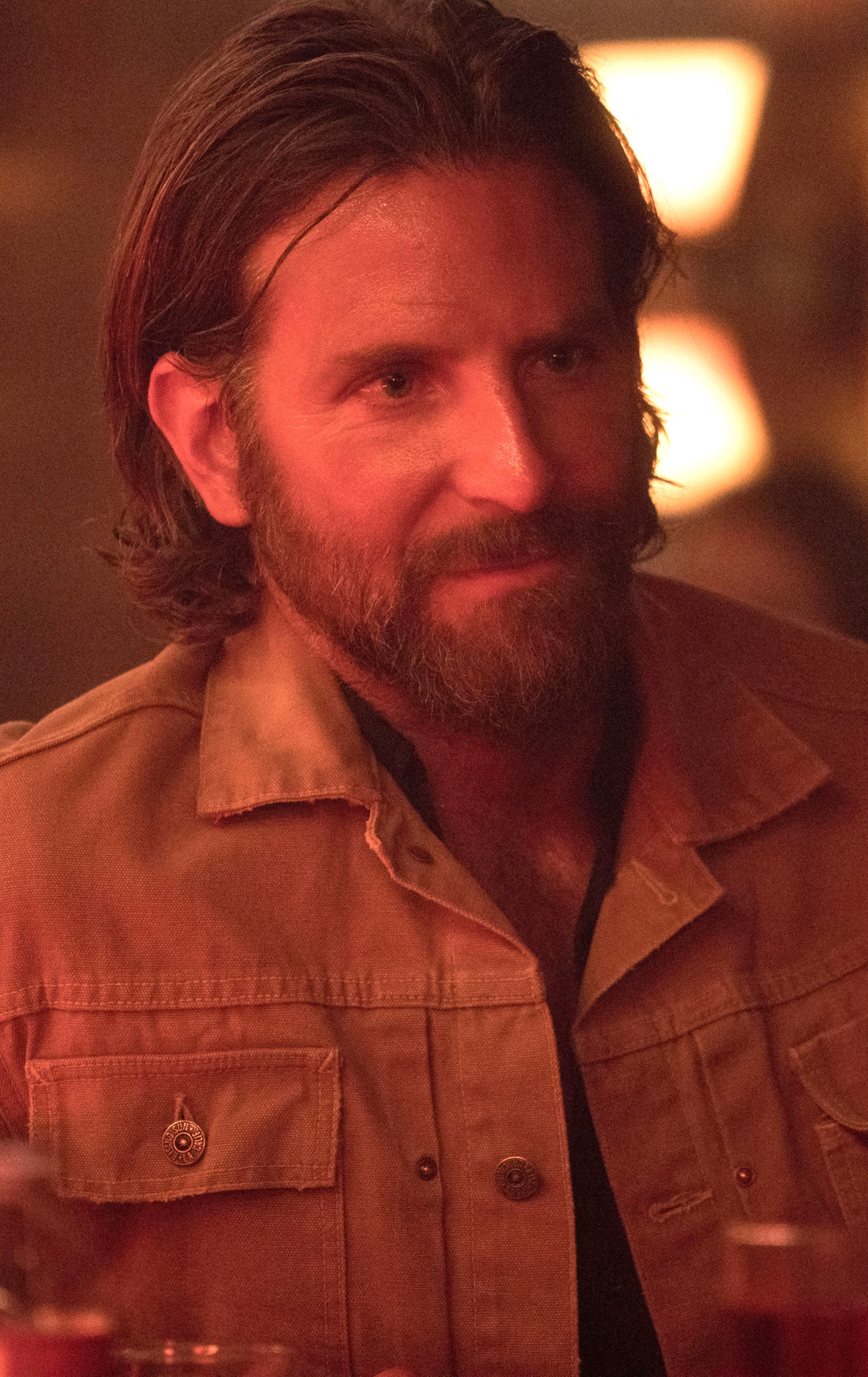 Bradley Cooper with long hair and a beard, wearing a rusty jacket in a bar