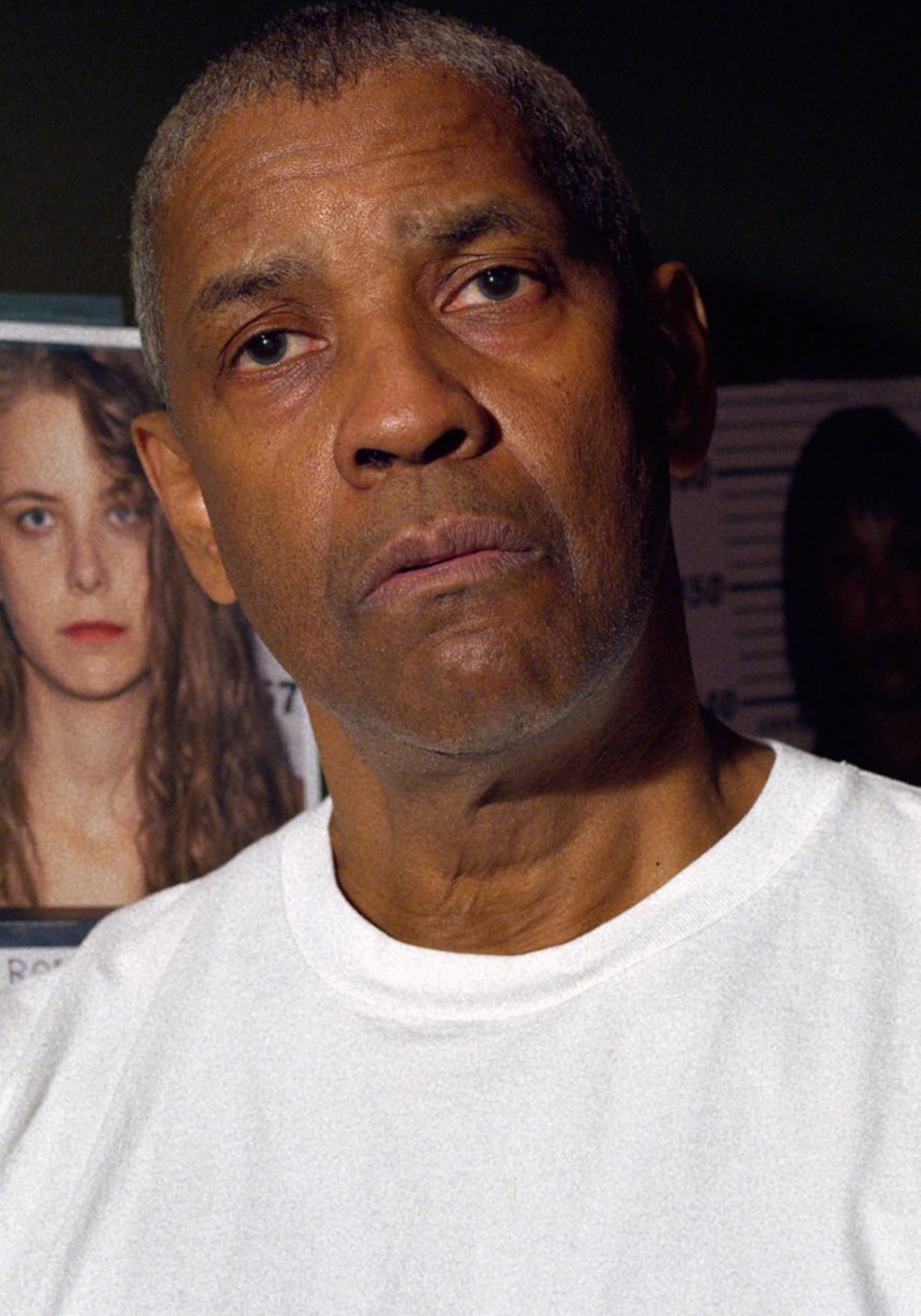 Denzel Washington wearing a white t-shirt while standing in front of images of potential suspects