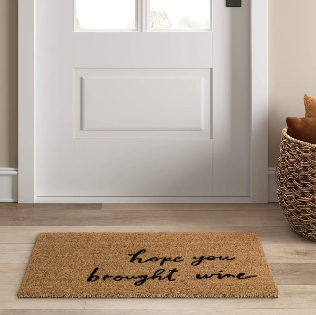 The light brown mat says &quot;hope you brought wine&quot; in a script font and is on the wooden floor in front of a white door