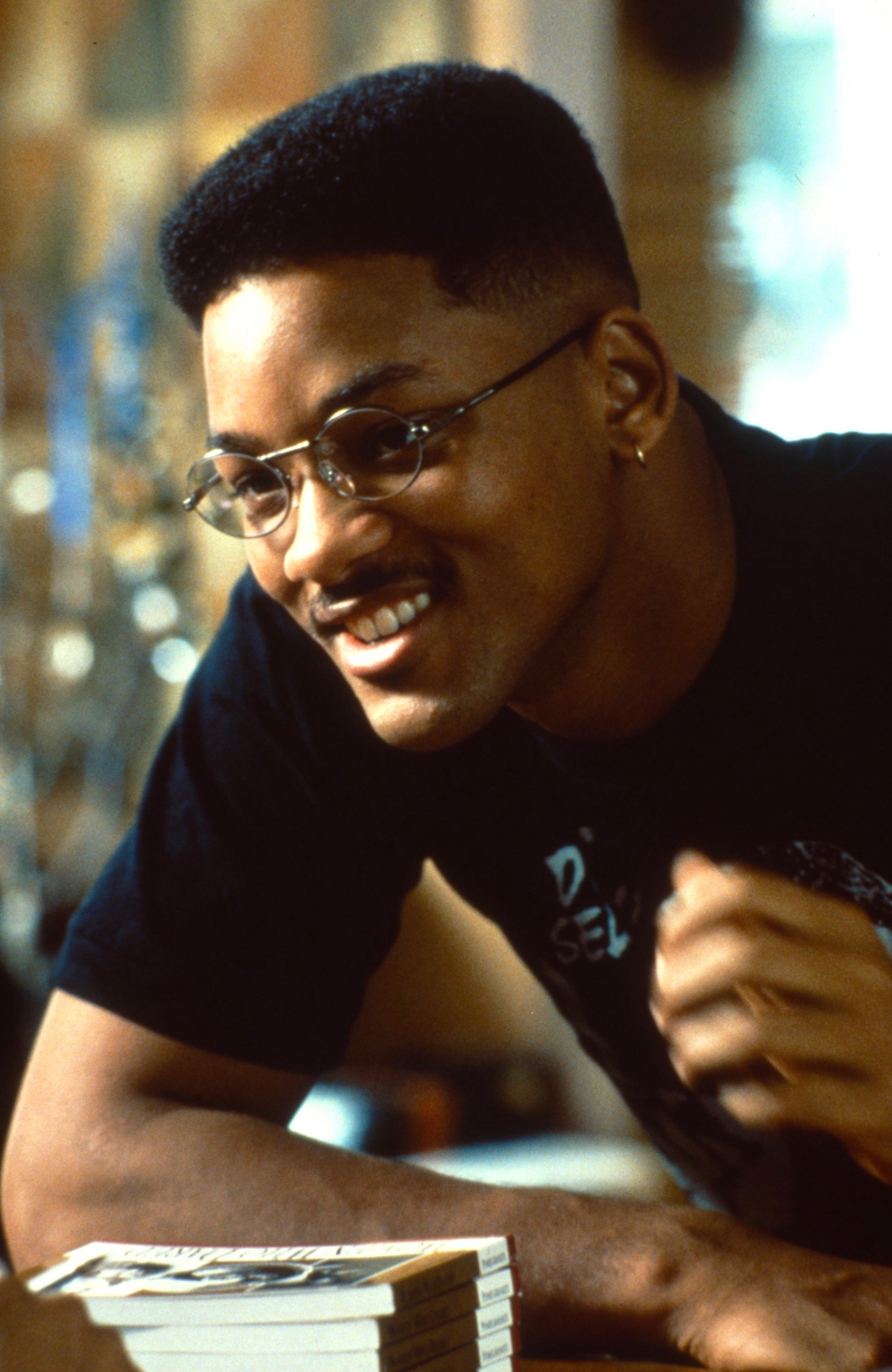 Will Smith wearing glasses, leaning on a table with a stack of books