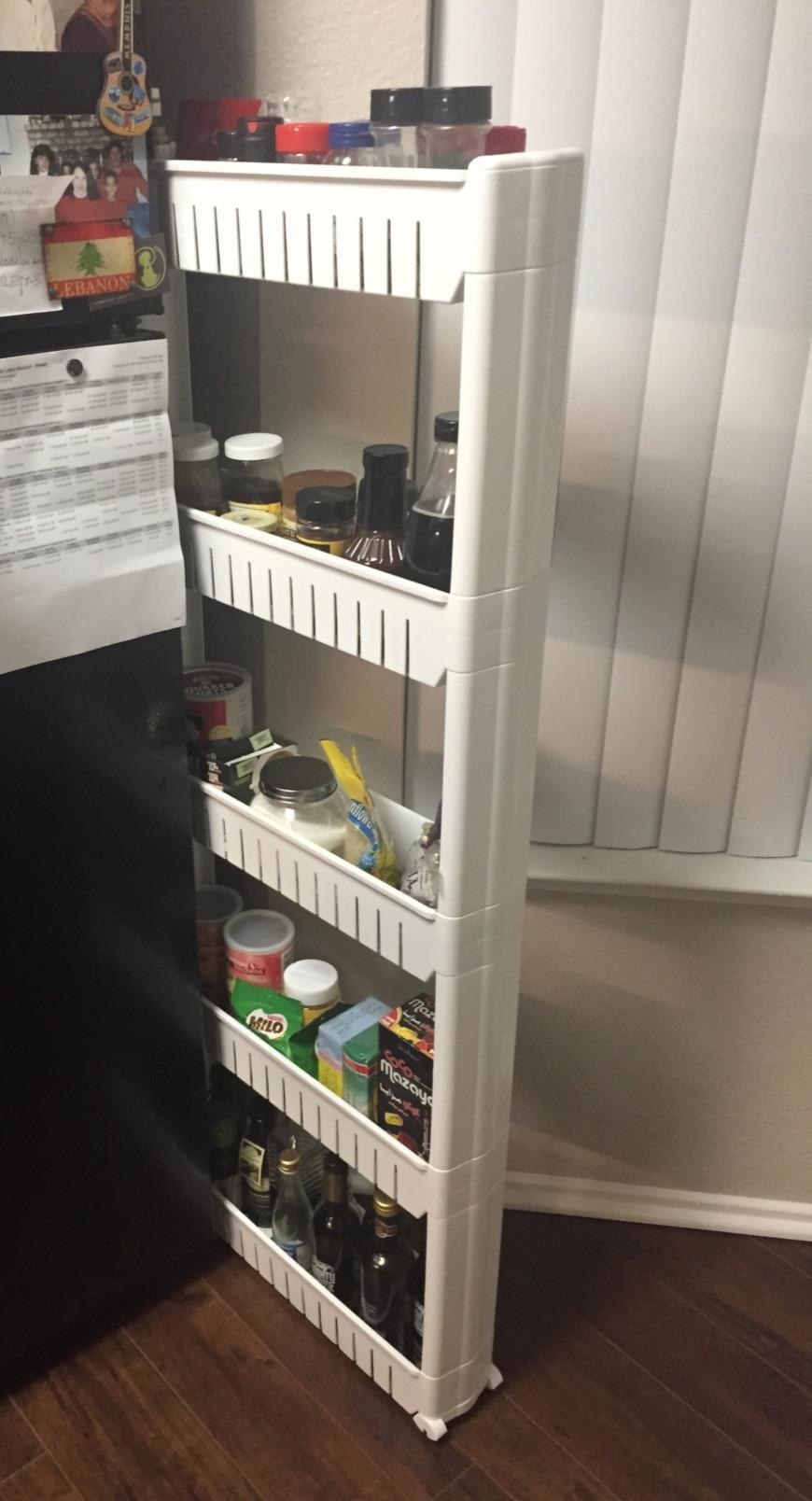 the rolling shelves with bottles, snacks, and cooking supplies on them, being stored between a fridge and a wall