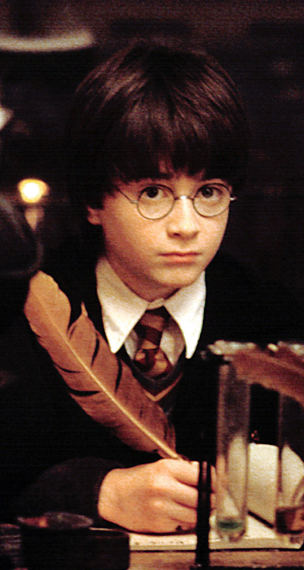 Daniel Radcliffe wearing round glasses while in a Hogwarts school uniform, writing