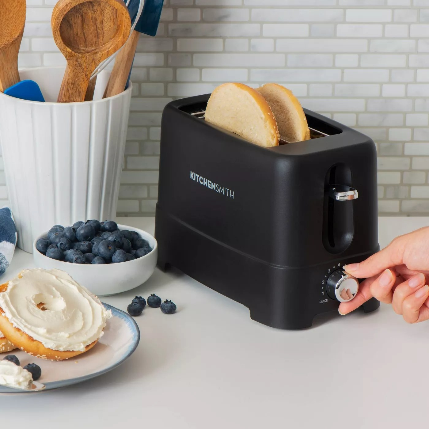 The black toaster says &quot;KITCHENSMITH&quot; and has two sesame bagel slices inside and is surrounded by blueberries, another bagel with cream cheese and various kitchen tools
