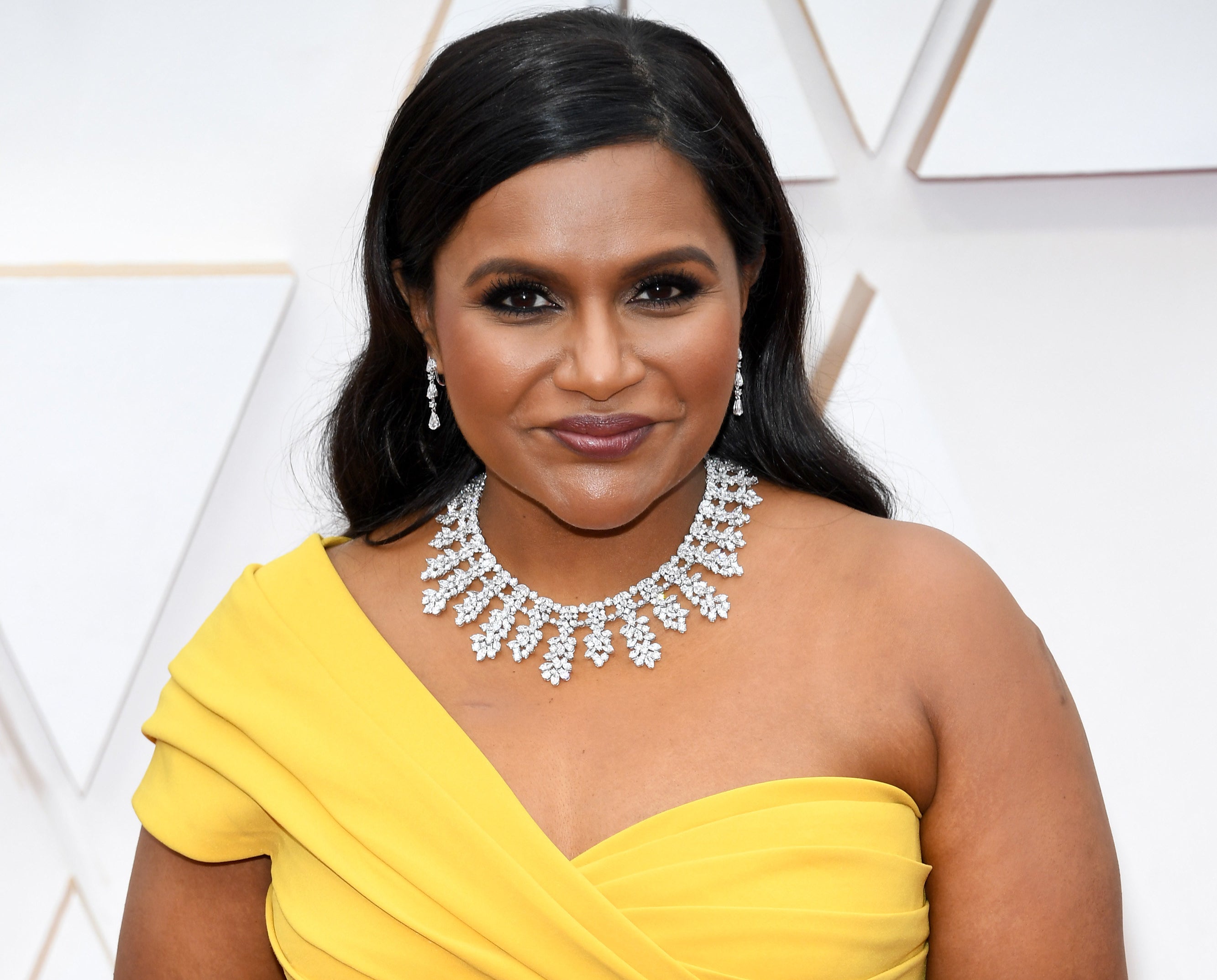 Mindy wears a yellow one-shoulder dress with a diamond necklace