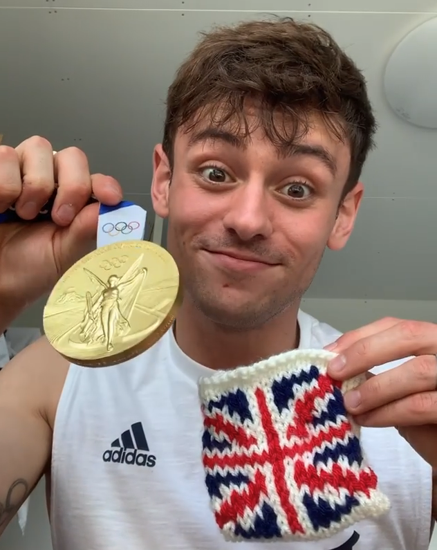 Tom smiling while holding up his Olympic gold medal and his knitted cosy