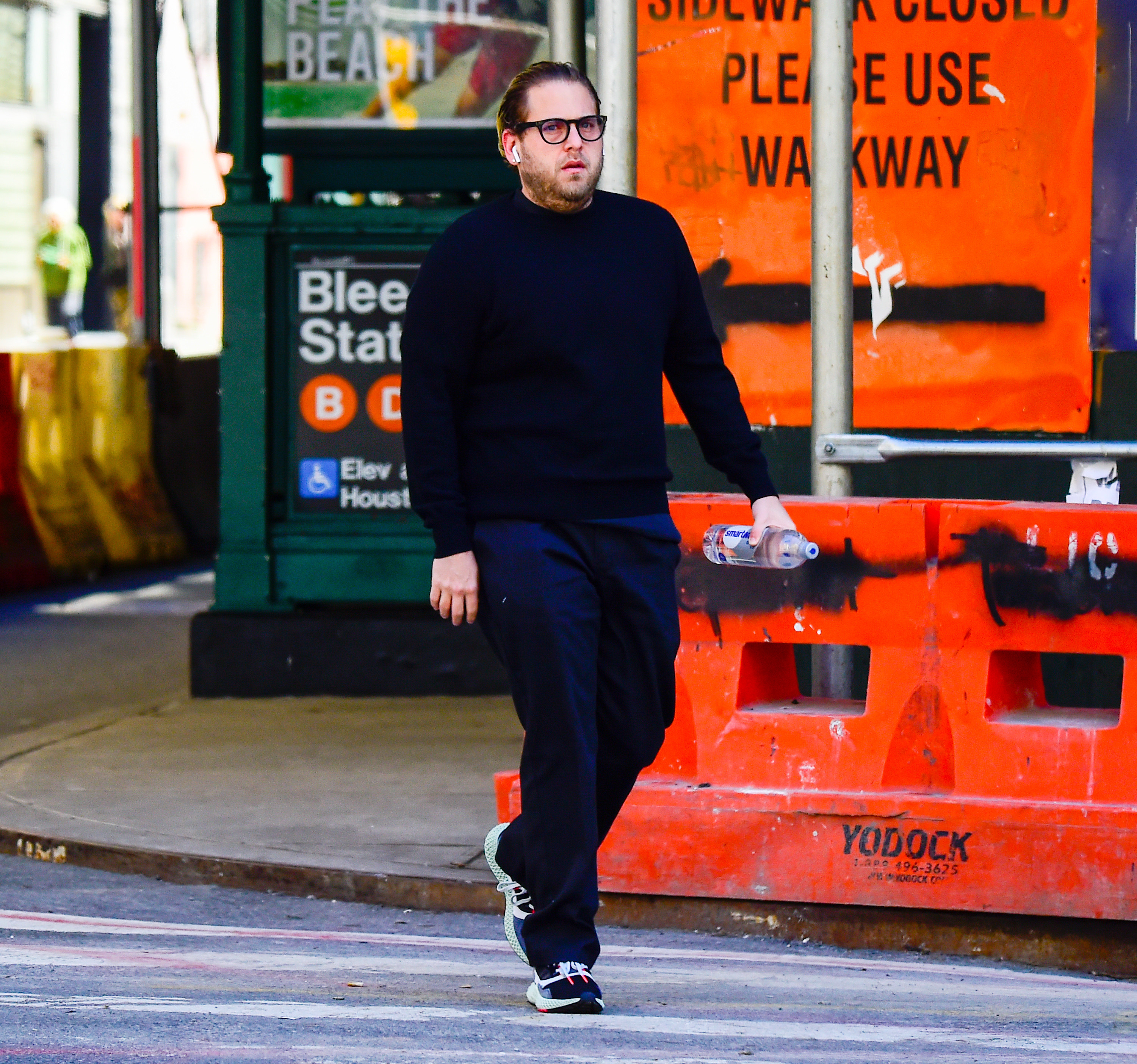 A bespectacled Jonah, wearing sneakers and holding a beverage bottle, walks past the Bleecker Street station in New York City