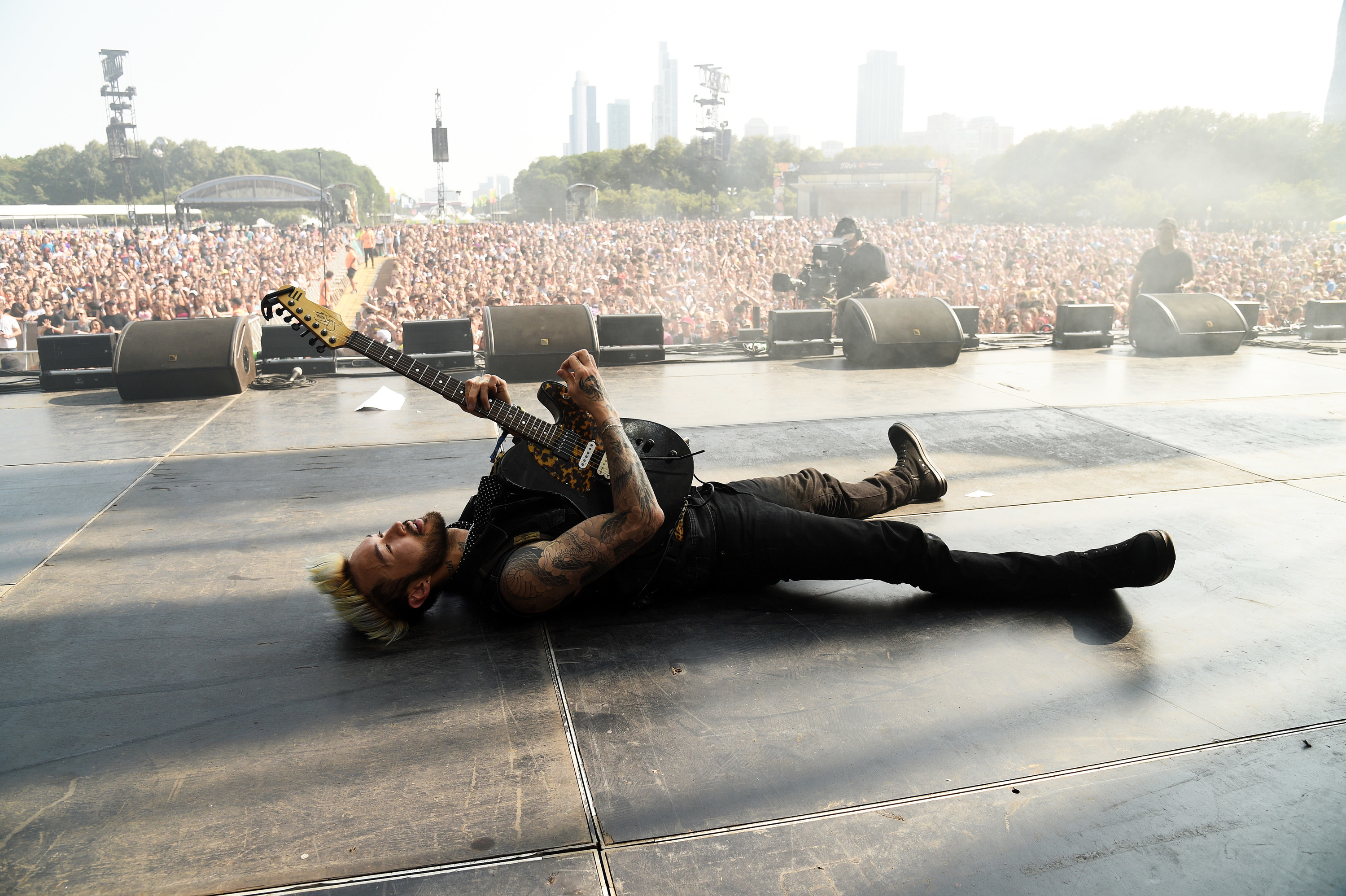 A musician is seen laying on stage holding a guitar as the crowd of thousands is seen in the background at Lollapalooza.