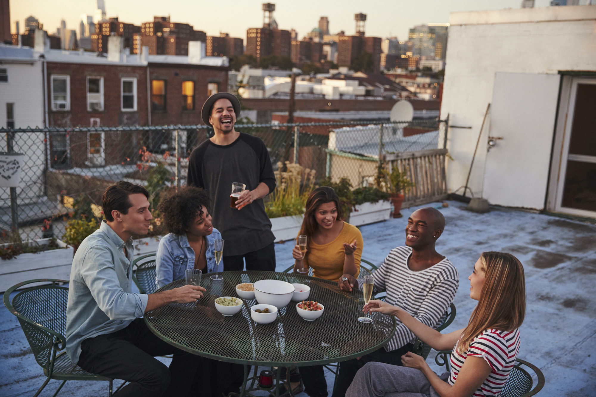 Friends in a city sit on a rooftop patio with drinks and snacks.