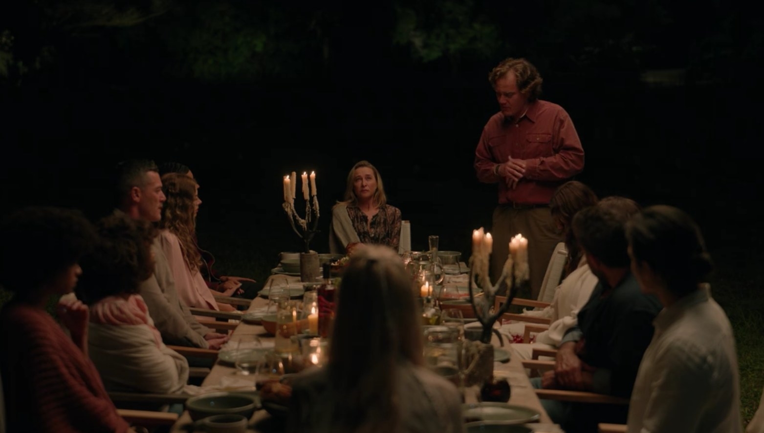 The guests sit around a dinner table outdoors at nighttime, Napoleon is standing to speak to them all