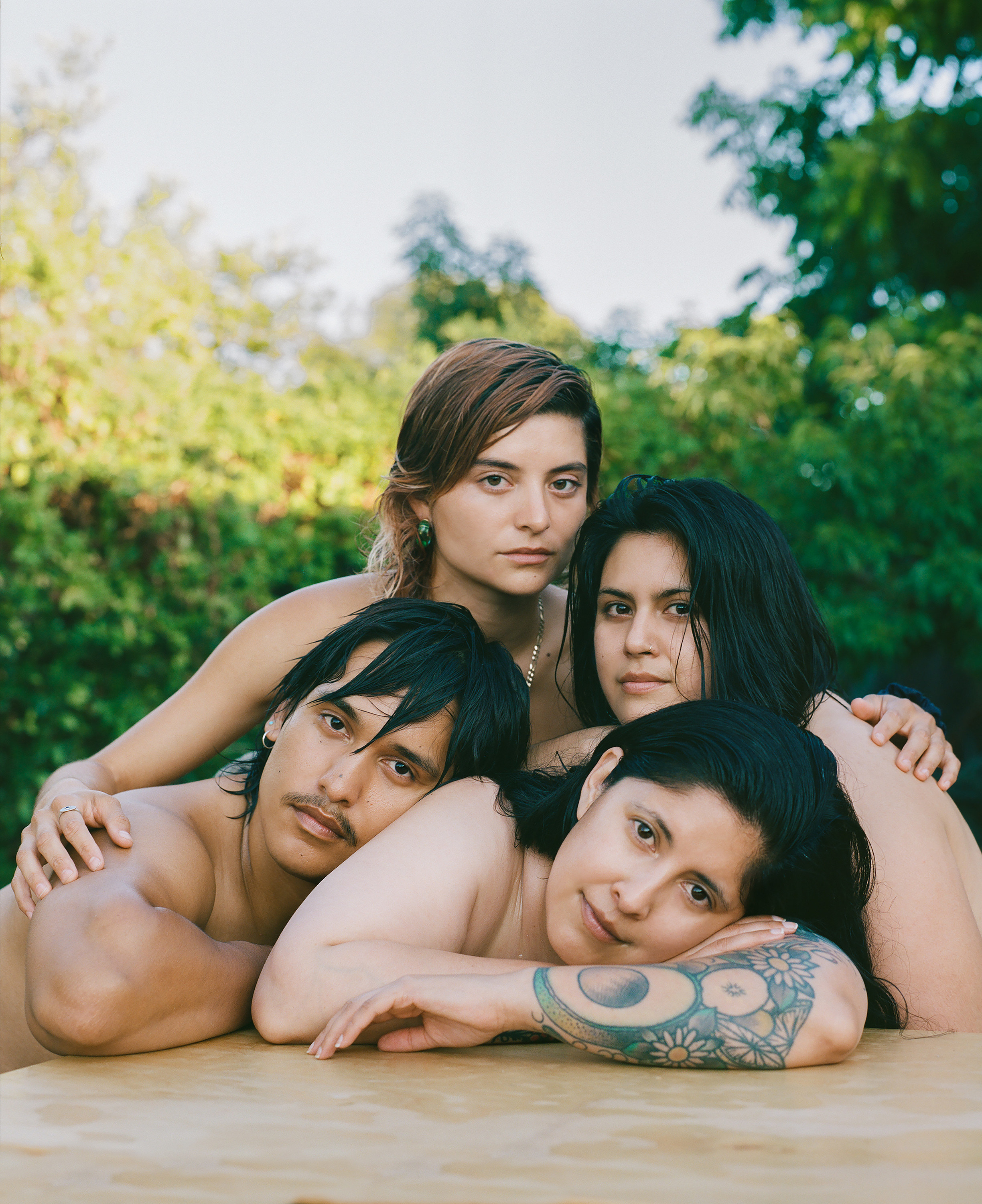 Four people, apparently naked, leaning on one another with their torsos on a plywood table