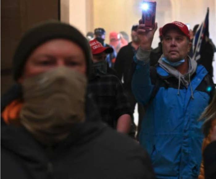 A crowd of people, with one masked man in the foreground and a man in a blue jacket holding up a phone