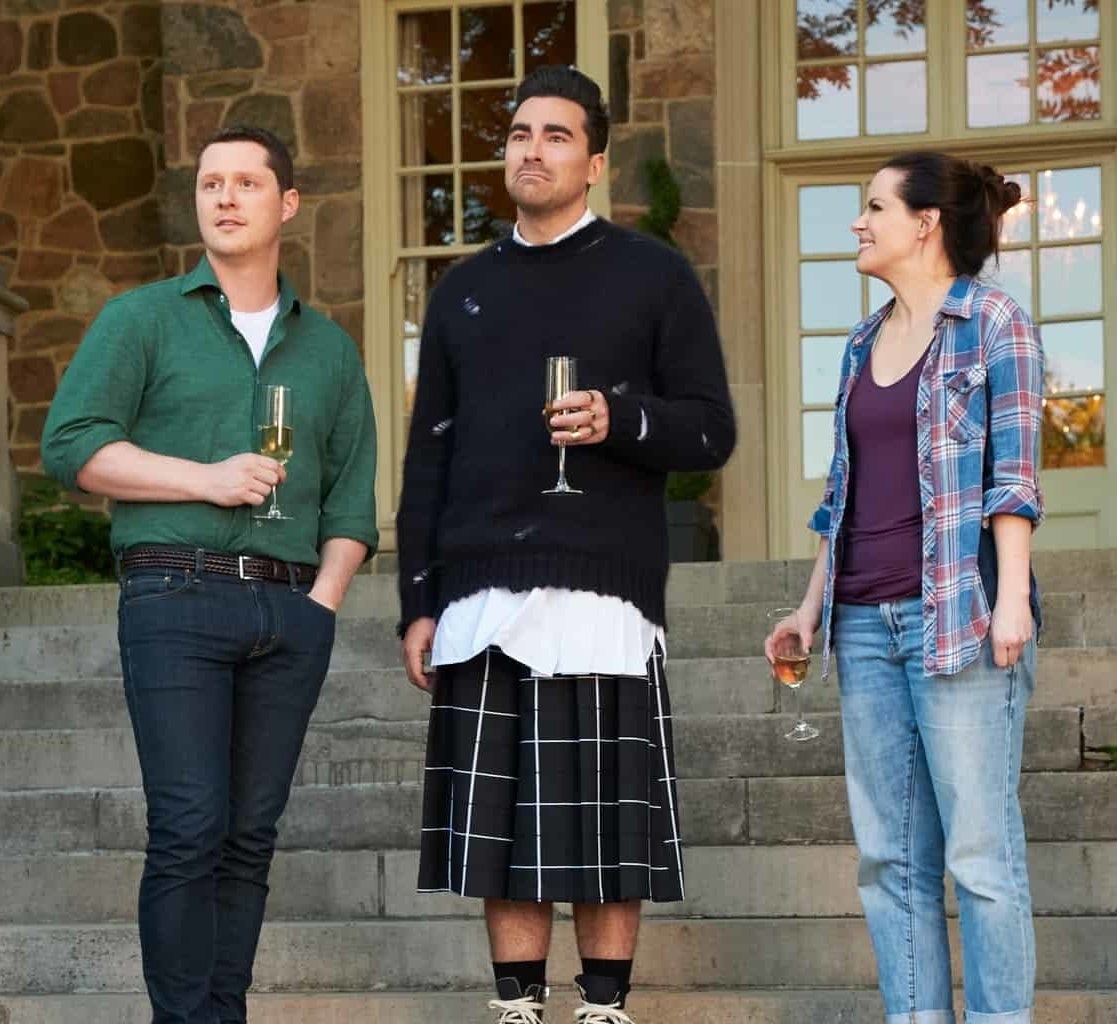 Patrick wearing a green button down shirt and dark jeans. David wearing a long black sweater over a long white shirt, a windowpane printed kilt. Stevie wearing a blue plaid shirt over a purple tank and jeans.