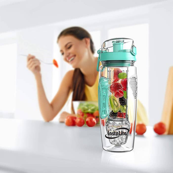 A transparent infuser bottle with fruits and a person enjoying a salad in the background