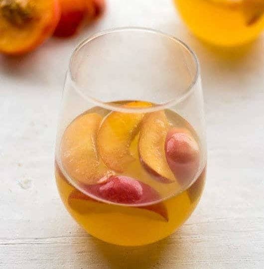 A stemless wineglass filled with a honey-colored white wine cocktail, garnished with peach slices