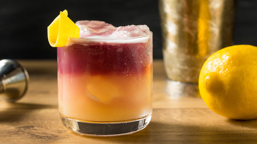 A layered cocktail in a tumbler: lemony whiskey sour on the bottom, red wine on the top, garnished with ice and a curl of lemon peel.