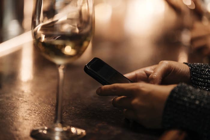 A glass of wine on a bar counter, with a person&#x27;s hands holding a phone
