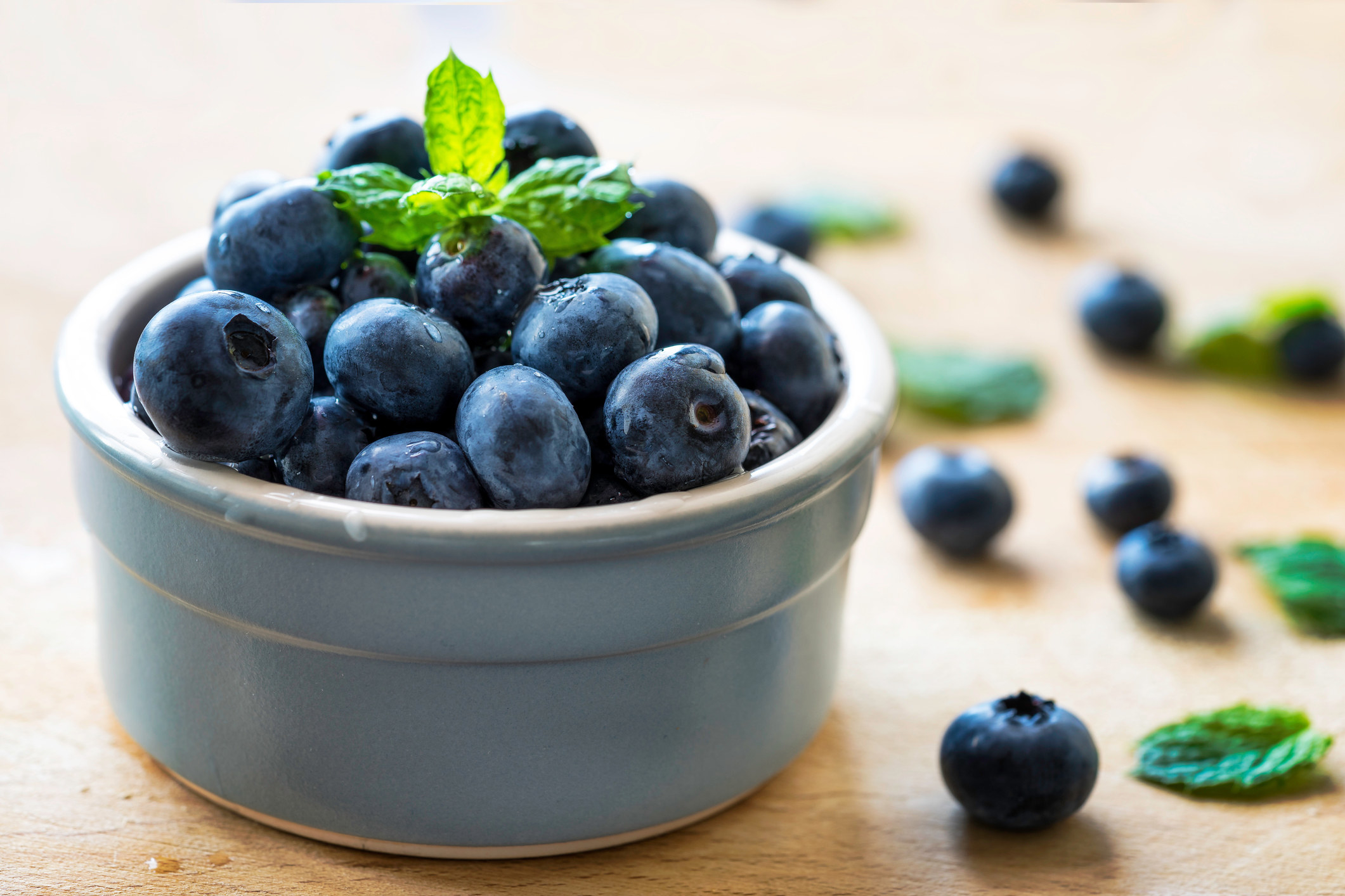A bowl holding blueberries, with other blueberries lying on the table around the bowl