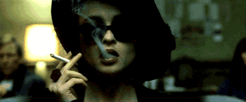 A shot zooms in on a woman wearing black sunglasses and a black hat smoking a cigarette.