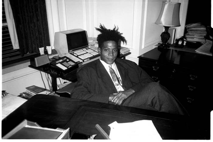 A black-and-white photo of Basquiat sitting at a desk in a suit and tie