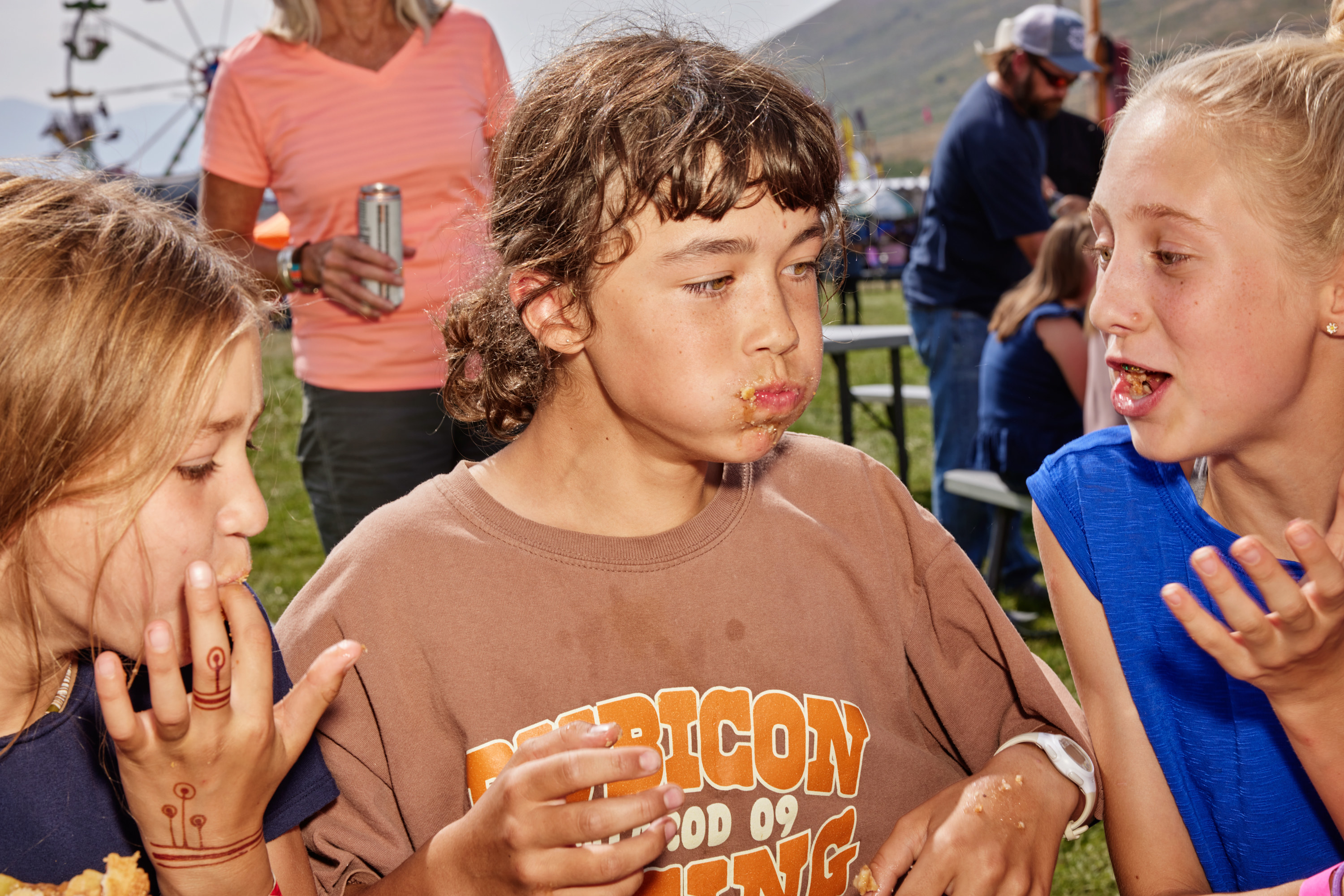 A girl, center, with her face stuffed full of pie as two girls sit on either side of her, also eating pie