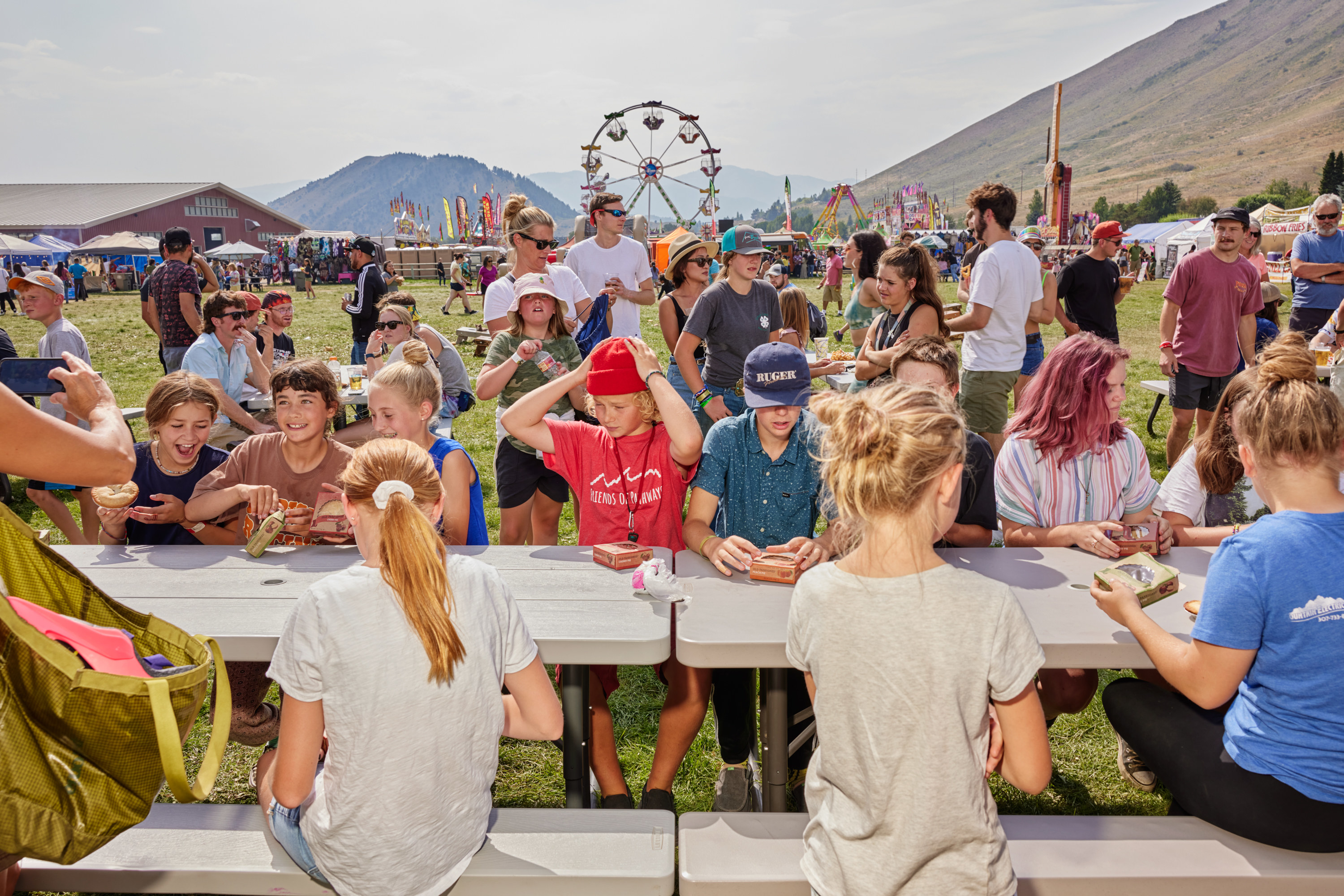 Children sitting at a picnic table with small pies, a ferris wheel, and parents in the background