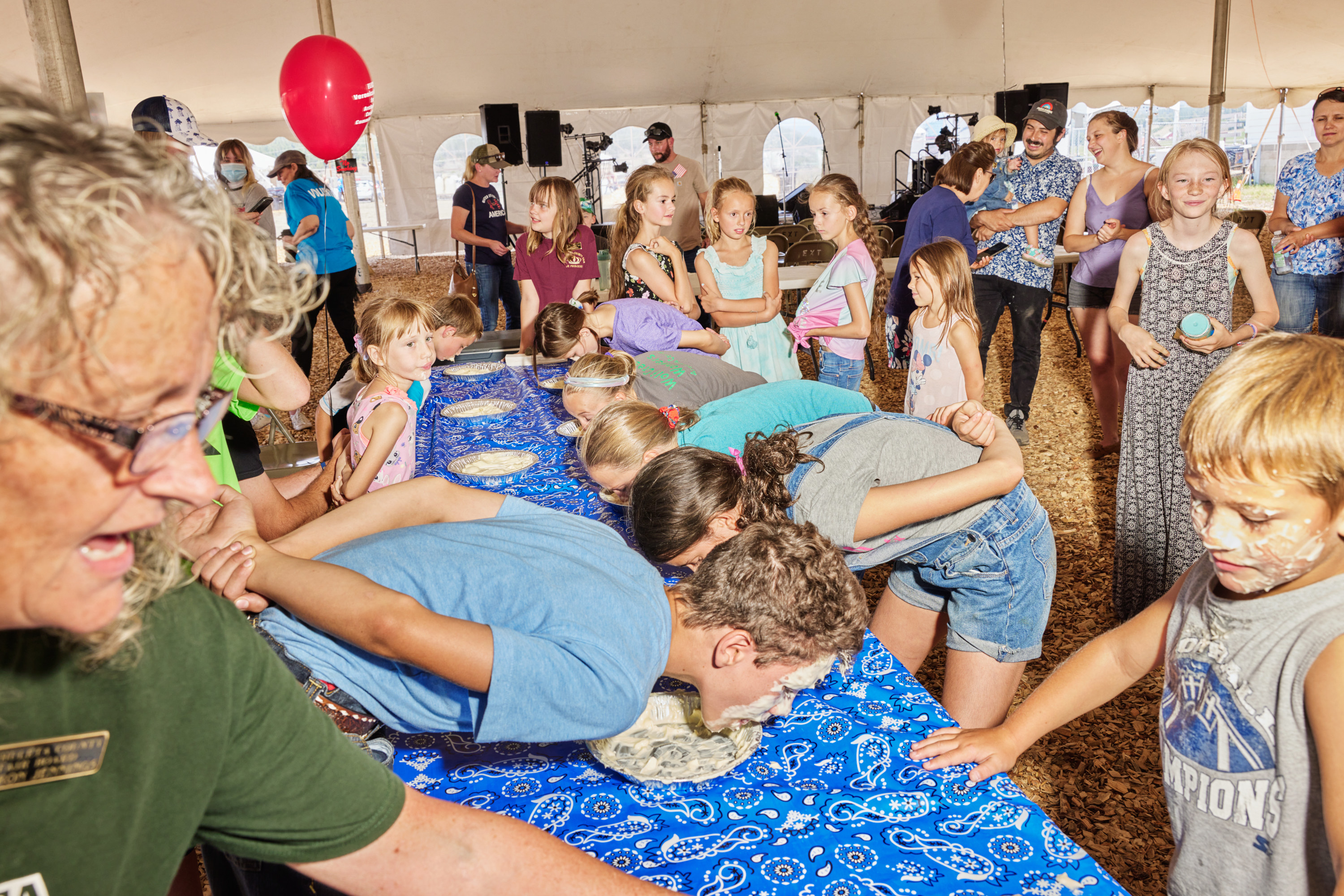 Two long rows of children with their heads bent down over a long table eating pie as a crowd looks on