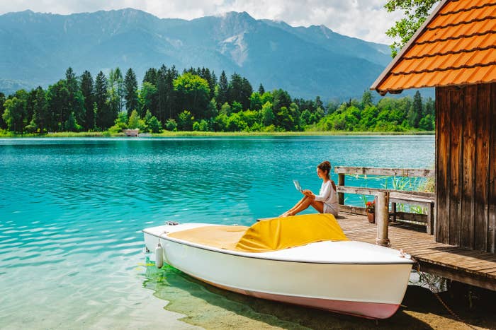 A woman sits at a water-front vacation house, near a boat working on her laptop