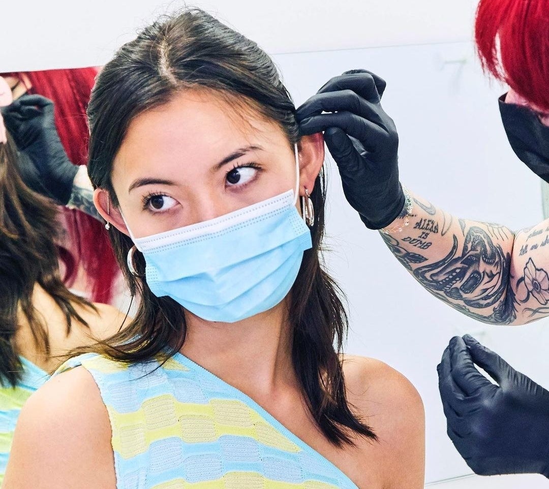 a model wearing a face mask having their ear pierced by a piercing technician