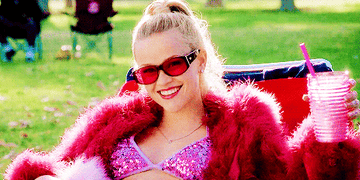 Elle smiles, holding a drink and wearing a bright pink ensemble.